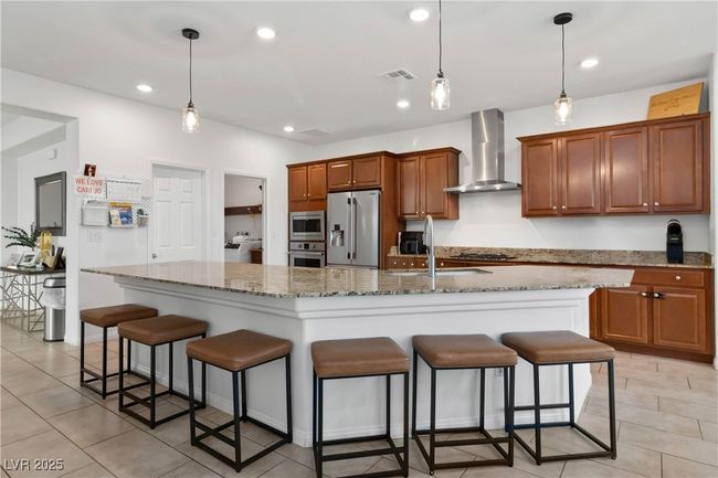 Kitchen featuring a breakfast bar, brown cabinets, stainless steel appliances, wall chimney range hood, and recessed lighting | Image 15