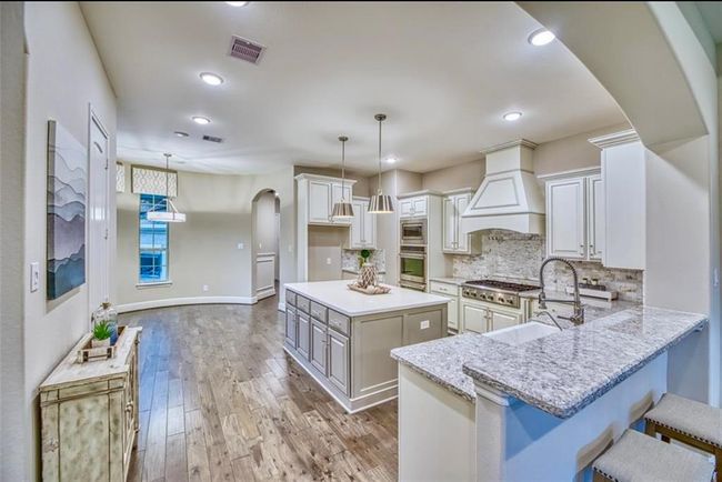 This kitchen features a spacious, open layout with a large island and breakfast bar. It has modern pendant lighting, granite countertops, and ample cabinetry. The hardwood floors and neutral color scheme add warmth and elegance. | Image 17