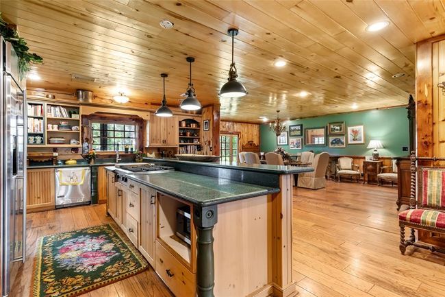 This photo showcases a warm, rustic kitchen and living area with wooden ceilings and floors. The kitchen features a large island with a granite countertop and pendant lighting, while the open layout flows into a cozy dining and seating area with comfortable chairs and decorative wall art. | Image 17