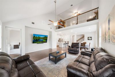 This image shows a spacious, well-lit living area with high ceilings, featuring a modern open-concept design that connects to a dining space and kitchen. The room is equipped with a ceiling fan and has a second-floor balcony overlook, providing a sense of openness and connectivity within the home. | Image 3