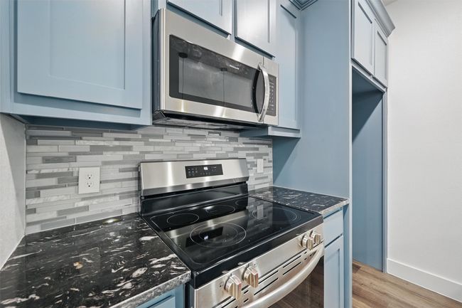 Kitchen featuring light wood-type flooring, dark stone countertops, blue cabinetry, decorative backsplash, and appliances with stainless steel finishes | Image 7