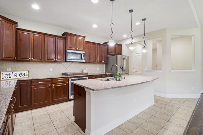 This bright kitchen features elegant cherry wood cabinetry, a large curved island with granite countertops, modern stainless steel appliances, and pendant lighting, offering a spacious and inviting area for cooking and entertaining. | Image 9