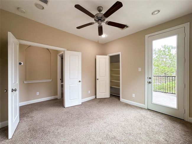 Alternate view of the room features neutral walls, plush carpet, and a ceiling fan. It includes a closet, a recessed shelf, and French doors leading to a private balcony with a view of trees. | Image 22