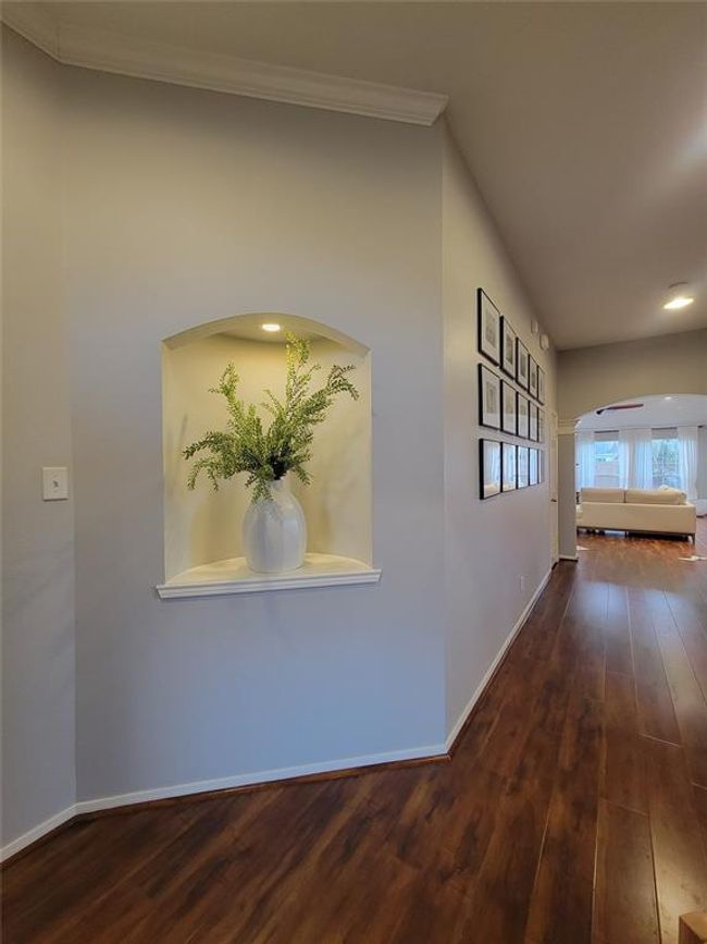 A welcoming hallway featuring elegant hardwood floors, recessed wall niche with decorative vase, and framed art. It leads into a bright, cozy living room with ample natural light. | Image 14