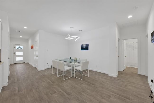 This photo showcases a modern, open-concept dining area with sleek wood flooring and white walls. It features a contemporary glass dining table with white chairs, and a unique light fixture above. The space is bright, with recessed lighting and natural light from the doorway. | Image 7