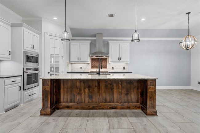 Kitchen featuring wall chimney exhaust hood, stainless steel appliances, white cabinets, decorative backsplash, and ornamental molding | Image 9