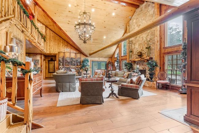 This spacious, rustic living room features a high vaulted ceiling with exposed wood beams, a large stone fireplace, and ample natural light from tall windows. The room is adorned with cozy seating and elegant decor, creating a warm and inviting atmosphere. | Image 23