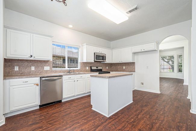 This kitchen features modern stainless steel appliances, white cabinetry, and a central island. The space is bright with natural light and has wood-style flooring, providing a warm and inviting atmosphere. An arched doorway leads to dining room, enhancing the open layout. | Image 7