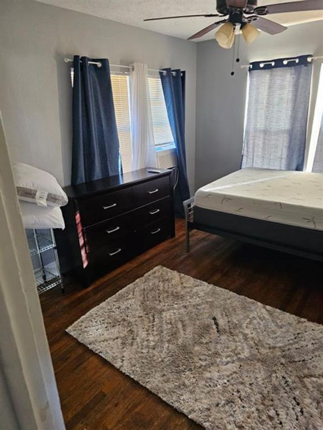 This cozy bedroom features hardwood floors, a ceiling fan, and ample natural light through multiple windows. It includes a comfortable bed, a dark wood dresser, and a soft area rug, creating a warm and inviting atmosphere. | Image 13