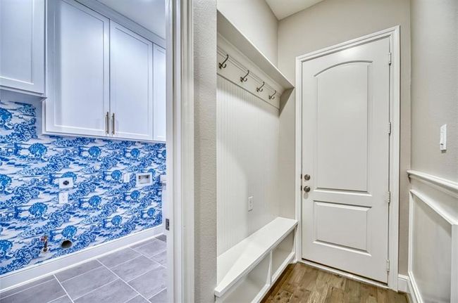 This photo shows a stylish mudroom with a bench and coat hooks, adjacent to a laundry area featuring striking blue and white patterned wallpaper, white cabinetry, and sleek gray flooring. | Image 34