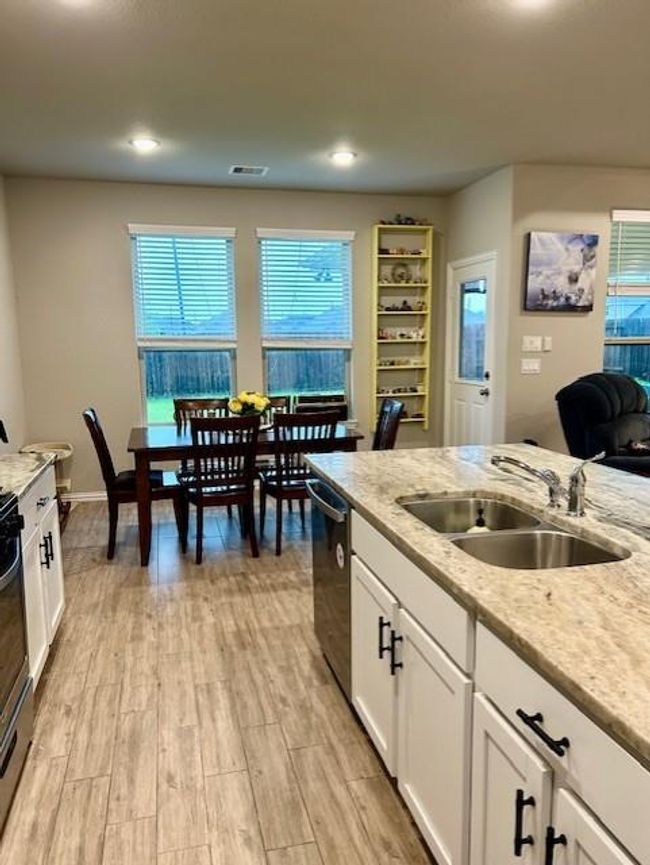 This photo showcases a modern, open-concept kitchen and dining area with light wood-look tile flooring and neutral walls. The kitchen features granite countertops, a double sink, stainless steel appliances, and white cabinetry. A dining table is positioned near large windows, providing natural light and a view of the backyard. A cozy sitting area is visible in the background, adding to the home's inviting atmosphere. | Image 12