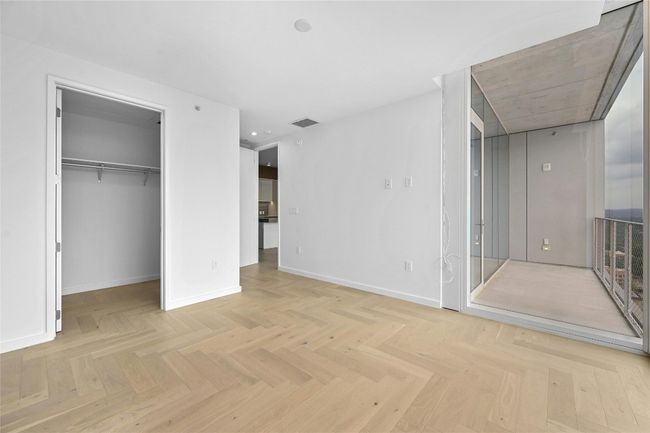The spacious primary bedroom features elegant light wood herringbone flooring and clean, modern lines throughout. Floor-to-ceiling window allowing for abundant natural light. The room includes a generous walk-in closet with built-in shelving and hanging space. | Image 8