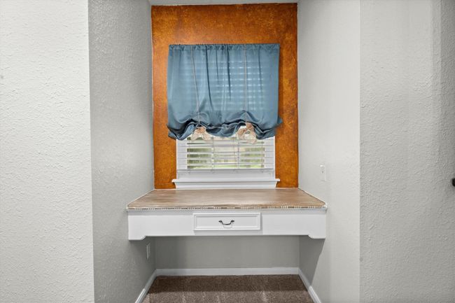 This photo features a cozy alcove with a built-in desk upstairs between secondary bedrooms, highlighted by a warm accent wall. A window with a blue curtain provides natural light, making it an ideal spot for a home office or study area. | Image 33