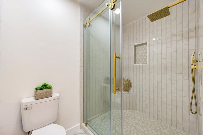 The primary bathroom features a luxurious walk-in shower, designed with modern elegance. The shower is equipped with a rain shower head, offering a spa-like experience with a gentle, soothing flow of water. The walls are beautifully finished with subway tile installed vertically, creating a sleek, contemporary look. The updated frameless glass door allows the design to shine, providing an open and airy feel. | Image 19