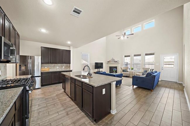 This open-concept living area features a modern kitchen with dark cabinetry, stainless steel appliances, and a large island. It flows into a spacious living room with high ceilings, abundant natural light, and a cozy seating area, perfect for family gatherings. | Image 4