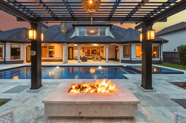 This stunning outdoor oasis delivers resort-style living at its finest. Centered around a luxurious pool and elevated infinity spa, the space is framed by a covered patio and expansive travertine deck—ideal for lounging or entertaining. A sleek fire pit beneath a contemporary pergola invites cozy evenings under the stars, while ambient lighting sets the mood. With seamless indoor-outdoor flow, this private retreat elevates both relaxation and entertaining in effortless style. | Image 41