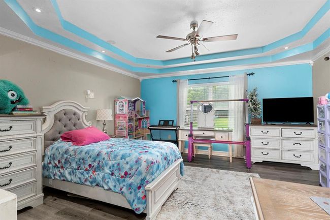 This is a spacious bedroom featuring a large bed with a tufted headboard, a dresser, a ceiling fan, and a TV stand. The room has bright blue walls with a unique light blue ceiling accent and is well-lit with natural light from a window dressed with sheer curtains. It also includes a play area with a dollhouse and a child's desk, suggesting it is currently styled for a child. The flooring is a light wood laminate. | Image 34