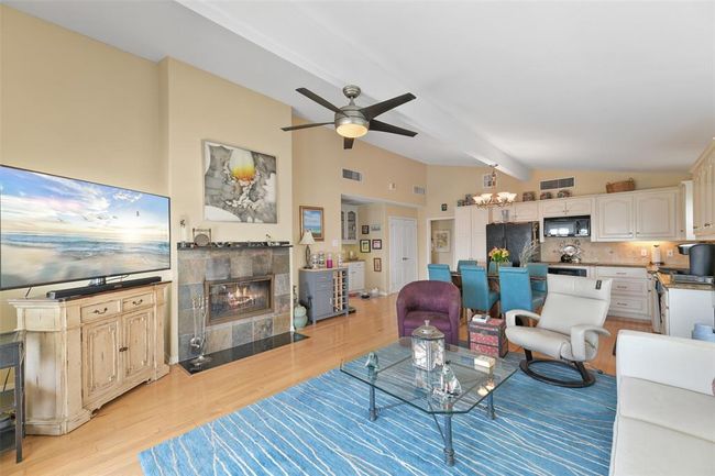 This bright, open-concept living area features high ceilings, a cozy fireplace, bamboo wood floors, and a stylish kitchen with white cabinetry. The space is enhanced by a large TV, contemporary furnishings, and a blue rug, offering a welcoming atmosphere perfect for relaxation and entertaining. | Image 13