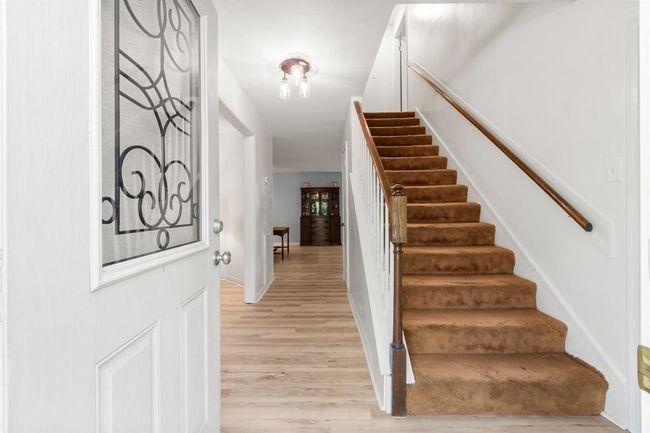 This photo showcases a bright entryway leading to a stairway with plush, tan carpeting. The area features light hardwood floors, a decorative glass-paneled door, and white walls, creating a clean and welcoming atmosphere. | Image 5