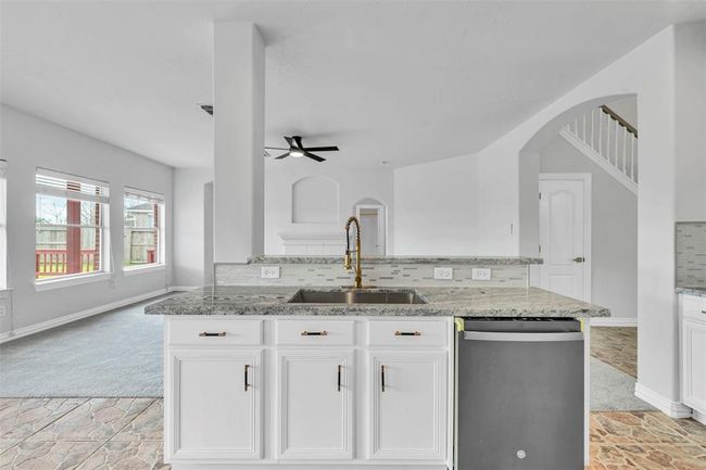 Bright and spacious kitchen with modern countertops, white cabinetry, and an open layout. It features a central island with a sink, overlooking a living area with large windows and a ceiling fan. Stairs lead up from the corner, adding an elegant touch. | Image 18