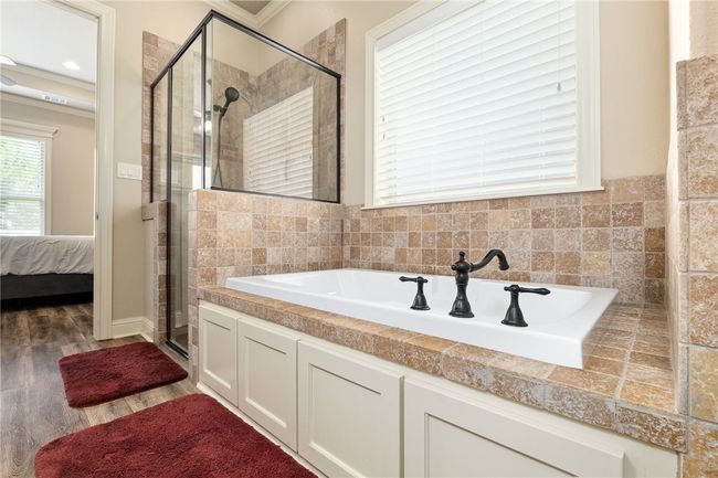 Across from the vanity, you'll find a glass-enclosed walk-in shower and a separate soaking tub. A large window with blinds, neutral tile design, and clean lines create a sleek, spa-like atmosphere. | Image 20