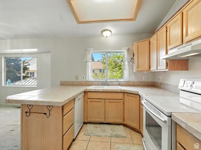 Kitchen featuring white appliances, under cabinet range hood, a peninsula, tile countertops, and light tile patterned flooring | Image 6