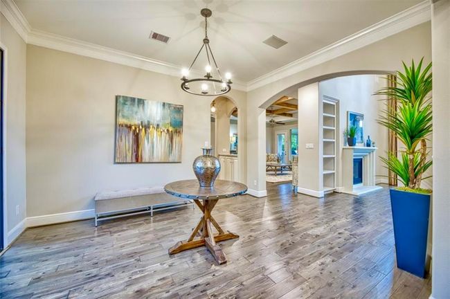This spacious entryway features elegant hardwood flooring, a stylish chandelier, and tasteful decor, including a round wooden table and artwork. The open archways lead to a bright living area with built-in shelving and a fireplace, creating a warm and inviting atmosphere. | Image 7
