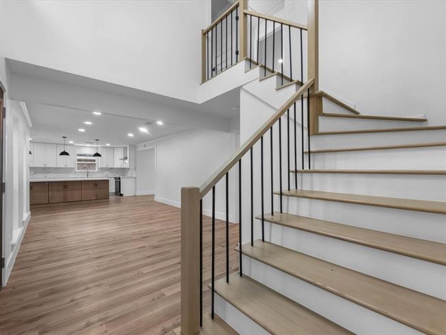 This photo showcases a modern, open-concept living space with sleek wooden flooring and a stylish staircase. The kitchen features elegant white cabinetry, marble countertops, and pendant lighting, creating a bright and inviting atmosphere. | Image 4