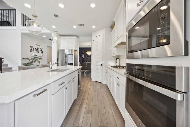 Modern kitchen with sleek white cabinetry, stainless steel appliances, and a spacious island with pendant lighting. The open layout leads to a dining area, featuring elegant wood flooring and ample natural light. | Image 17