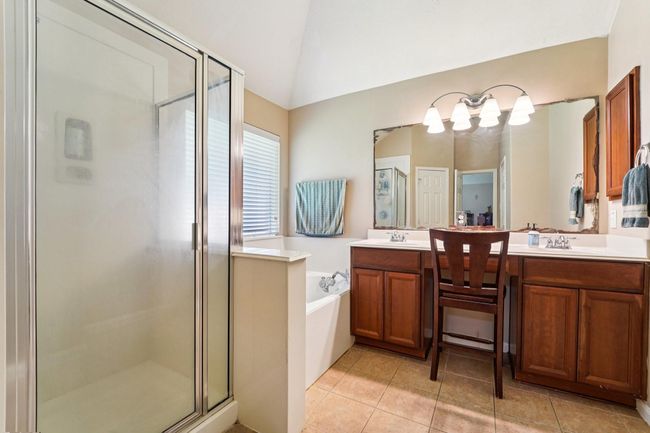 This bathroom features a spacious glass-enclosed shower, a large vanity with dual sinks, and warm wood cabinetry. Natural light fills the room through a window near the bathtub, creating a bright and inviting space. | Image 15