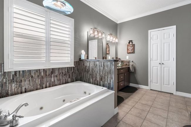 This photo features a modern bathroom with a spacious Jacuzzi tub and stylish tile backsplash. It includes a double sink vanity with elegant lighting and ample storage. The room is enhanced by neutral tones and natural light from large windows with shutters. | Image 17