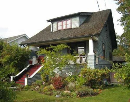 Craftsman-style home featuring a prominent dormer, front porch with white railing, red-painted exterior stairs, mixed siding with shingles, and extensive mature landscaping | Card Image