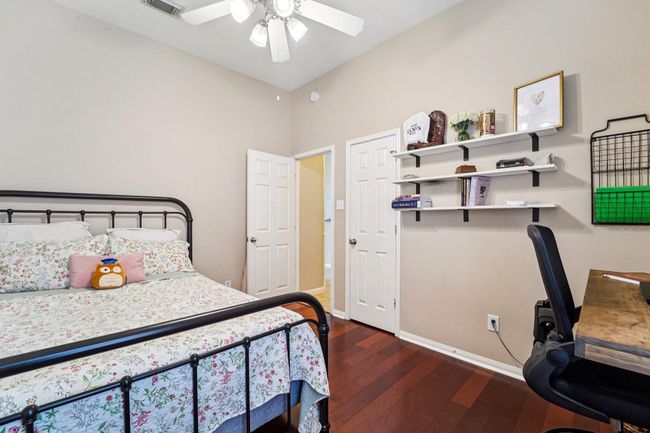 Cozy bedroom with warm wood flooring, featuring a decorative metal bed and floral bedding. The room includes built-in shelves, a small desk area, and a ceiling fan for comfort. Neutral walls enhance the inviting atmosphere. | Image 20