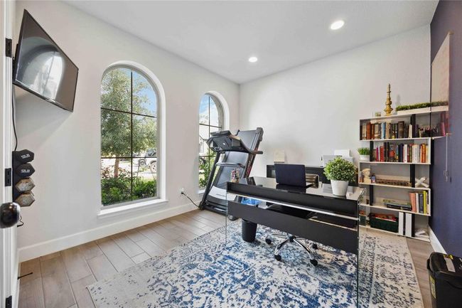This home office features large arched windows that provide ample natural light. It includes a treadmill, a sleek desk with a laptop, and a bookshelf, creating a perfect blend of workspace and exercise area. The room is bright and inviting, with stylish decor and a cozy atmosphere. | Image 5