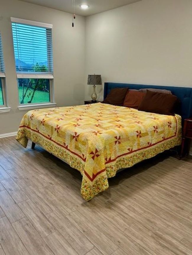 This bedroom features a cozy setup with a large bed adorned with a colorful quilt. Natural light filters in through two windows, and the room has a modern wood-look tile floor. A bedside lamp adds a touch of warmth. | Image 13