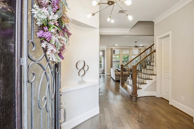 This inviting entryway features a decorative wrought iron door. It leads to a bright living area with large windows, a modern light fixture, and a staircase with wooden railings. The space has hardwood floors and is open and welcoming. | Image 5