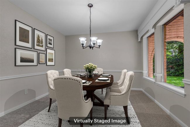 This is a virtually staged dining room featuring a classic round table with upholstered chairs. The room is well-lit by a stylish chandelier and has large windows offering a view of the greenery outside. Neutral walls are accented with framed artwork, creating a cozy and inviting atmosphere. | Image 10