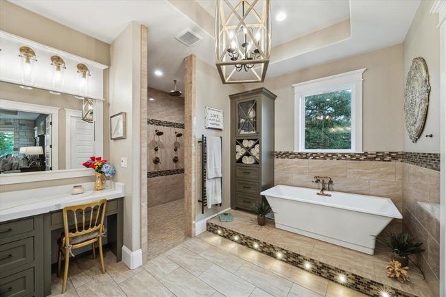 The primary bathroom features an oversized soaker tub set on an elevated light platform, creating a spa-like atmosphere. A high window allows for abundant natural light, while the custom ceramic tile design behind the tub complements the lighted platform, adding a touch of elegance and symmetry to the space. | Image 18
