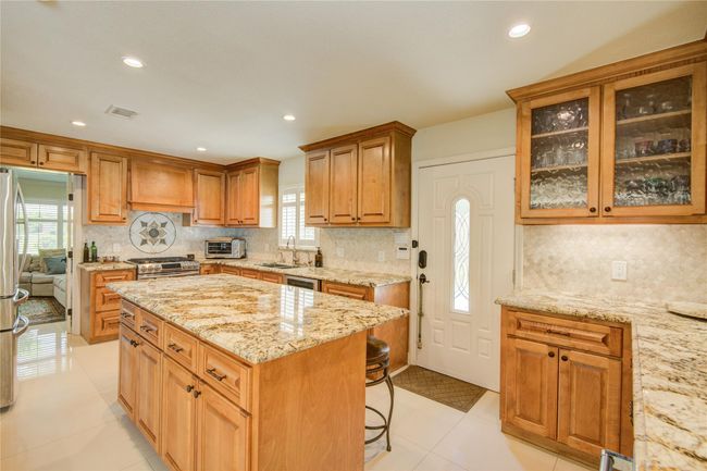 The kitchen offers abundant soft-close wood cabinetry, stainless steel appliances, and a spacious granite island with seating, plus a peek into the formal living room to the left. | Image 17