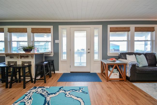 This inviting living space features large windows and a glass pane door providing abundant natural light. It includes a seating area with a sofa, a stylish area rug, and a breakfast island with stools, offering a blend of comfort and functionality for entertaining. | Image 12