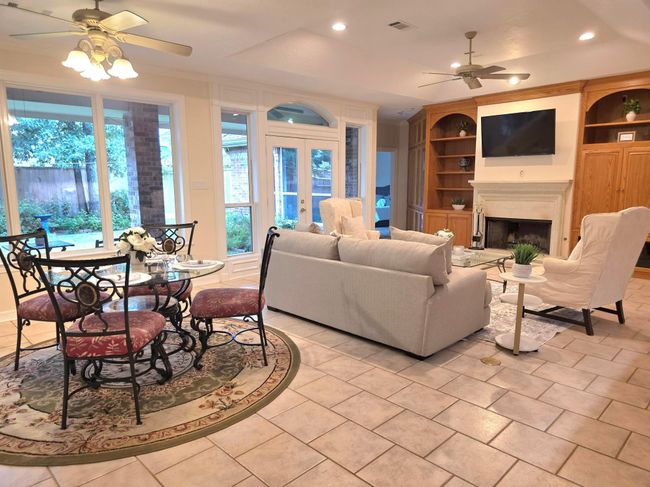 This inviting living area features a cozy fireplace, built-in wooden shelves, and ample natural light from large windows. The open layout includes a dining space with elegant seating, perfect for entertaining. Neutral tones and tile flooring add to the room's charm and functionality. | Image 22