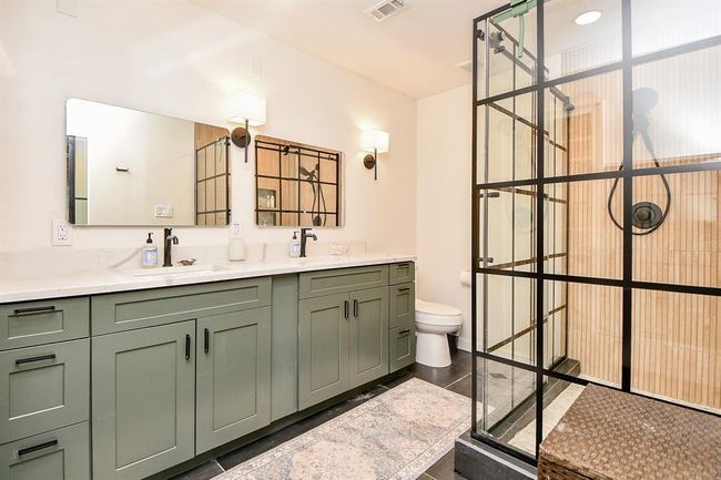 This bathroom features a modern, spacious design with green cabinetry, dual sinks, and sleek black fixtures. The large glass shower enclosure adds a touch of elegance, complemented by ample lighting and a neutral color palette. | Image 4