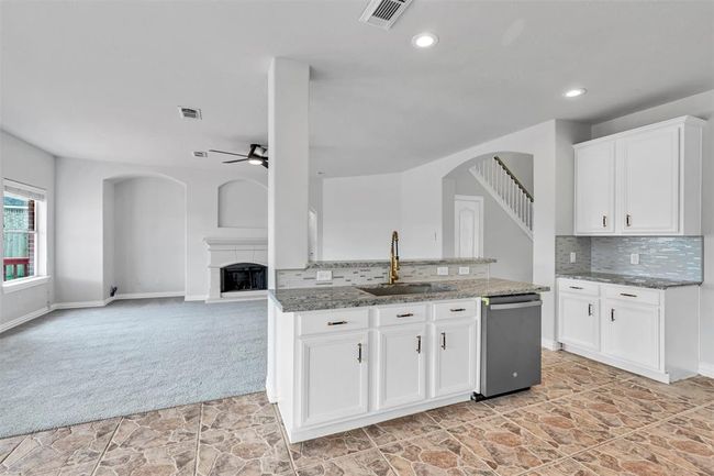 This photo showcases a bright, open-concept living area with a modern kitchen featuring granite countertops, white cabinetry, and stylish tile flooring. The adjoining living room has a cozy carpeted area, large windows for natural light, and a fireplace, with stairs leading to the upper level. | Image 15