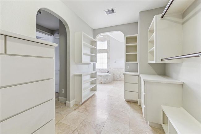 A spacious, well-lit walk-in closet with ample shelving and storage. It features an arched doorway leading to a luxurious bathroom with a large window and a soaking tub. | Image 19