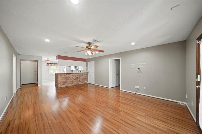 This photo showcases a spacious open-concept living area with vinyl wood flooring. A ceiling fan is centrally located, and the room connects to a kitchen with a brick accent and modern appliances, offering a welcoming and versatile space. | Image 5