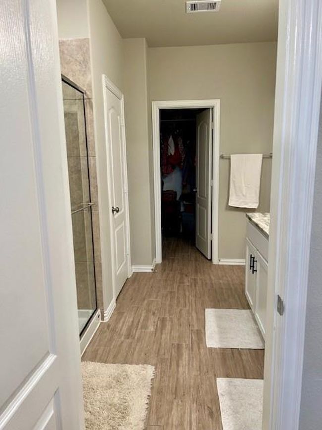 This photo shows a modern bathroom with wood-look tile flooring, a glass-enclosed shower on the left, and a vanity with a granite countertop on the right. It leads to a walk-in closet at the end. | Image 18