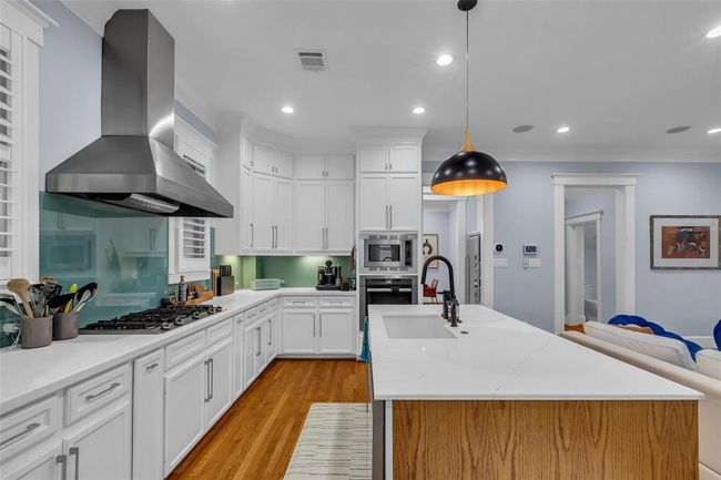 This modern kitchen features sleek white cabinetry, a central island with a sink, stainless steel appliances, and a gas cooktop. The space is well-lit with recessed lighting and a stylish pendant lamp, complemented by hardwood floors and a soft grey wall color. | Image 16
