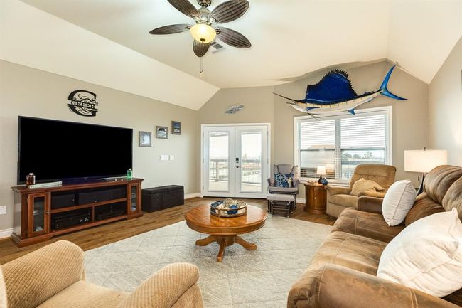 Spacious living room with vaulted ceiling, featuring cozy seating, large TV, ceiling fan, and natural light from french doors and windows. Decor includes nautical elements and wood flooring. | Image 4