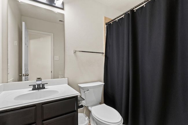 This bathroom features a sleek design with a modern vanity, a large mirror, and a clean white sink. It includes a toilet and a shower area with a black curtain, offering a stylish contrast against the neutral walls. | Image 25