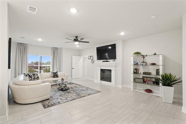 Family living room with light wood flooring and neutral tones. Features include a fireplace with a mounted TV, large window for natural light, and ceiling fan | Image 6